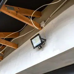 Nowoczesna instalacja elektryczna w garażu: jasny reflektor LED z czujnikiem ruchu zamontowany na betonowej belce, widoczne drewniane krokwie i białe przewody.