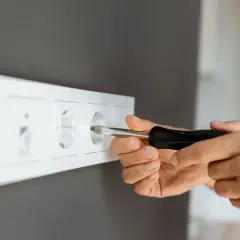 Ręce majsterkowicza przykręcają gniazdko, pokazując, jak przenieść gniazdko bez kucia.