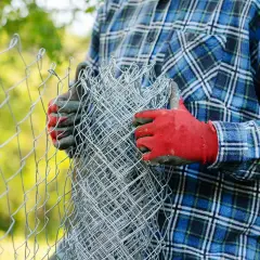 Osoba w czerwonych rękawicach i kratowanej koszuli przygotowuje się do montażu siatki ogrodzeniowej, trzymając zwinięty materiał.