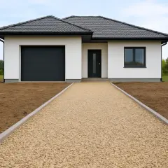 Nowy dom z garażem i drzwiami wejściowymi. Przed nim rozciąga się podjazd z kruszywa, otoczony trawnikiem.