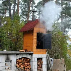 Prosta wędzarnia z cegły, z której unosi się dym. W środku wiszą kiełbaski. Obok stos drewna.