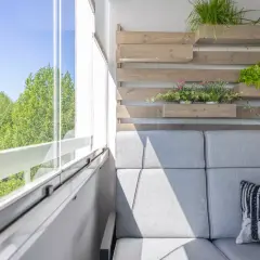 Przytulny mały balkon w bloku z szarą sofą, poduszką z liskiem i zielenią na drewnianej ściance.
