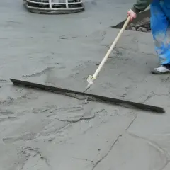Pracownik w niebieskich ogrodniczkach wygładza świeży beton za pomocą łaty. Trwa wygrzewanie posadzki.