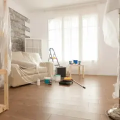 Pokój przygotowany do malowania paneli podłogowych. Meble i lampa przykryte folią, na podłodze wałek i farby.