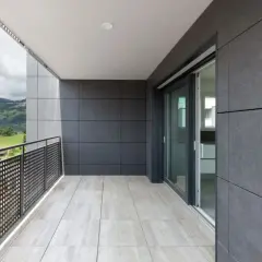 Nowoczesny balkon z jasnymi płytkami na podłodze i szarymi płytkami na ścianach. Widok na zielone wzgórza i budynki.