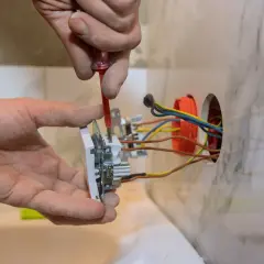 Ręce elektryka dokonują montażu gniazdka, przykręcając je do ściany. Widać kolorowe przewody wychodzące z puszki instalacyjnej.