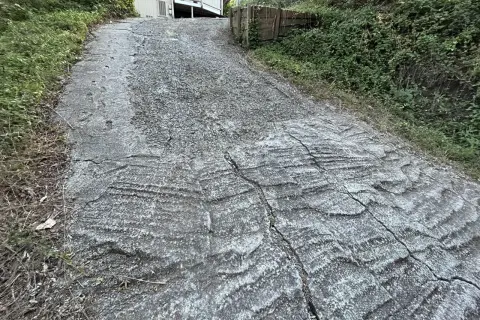 Wyboisty, wysoki podjazd do garażu, z widocznymi pęknięciami i nierównościami, otoczony zielenią.