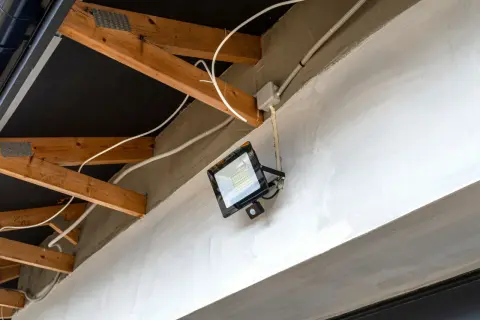Nowoczesna instalacja elektryczna w garażu: jasny reflektor LED z czujnikiem ruchu zamontowany na betonowej belce, widoczne drewniane krokwie i białe przewody.
