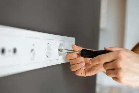 Ręce majsterkowicza przykręcają gniazdko, pokazując, jak przenieść gniazdko bez kucia.