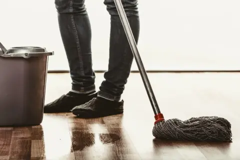 Osoba w jeansach i kapciach myje podłogę mopem, obok stoi wiadro. Naturalne czyszczenie octem.