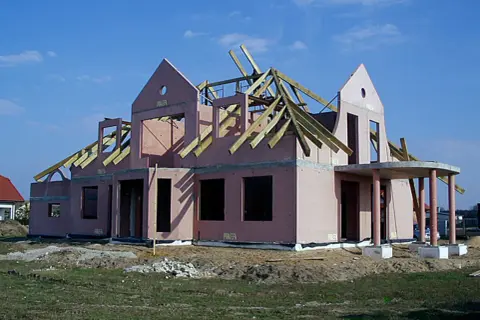 Budowa domu keramzytowego w różowym tynku, z widoczną konstrukcją dachu i wejściem z kolumnami.