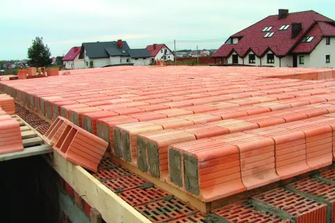 Strop ackermana w budowie, rzędy pustaków ceramicznych czekają na zalanie betonem. W tle domy z czerwonymi dachami.