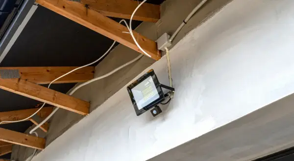 Nowoczesna instalacja elektryczna w garażu: jasny reflektor LED z czujnikiem ruchu zamontowany na betonowej belce, widoczne drewniane krokwie i białe przewody.