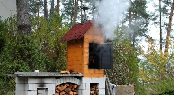 Prosta wędzarnia z cegły, z której unosi się dym. W środku wiszą kiełbaski. Obok stos drewna.