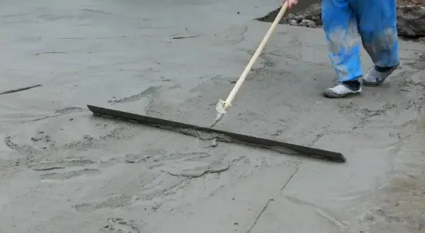 Pracownik w niebieskich ogrodniczkach wygładza świeży beton za pomocą łaty. Trwa wygrzewanie posadzki.