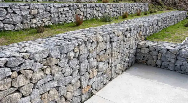Schody terenowe z kamiennych gabionów to świetny pomysł na murek oporowy. Widać kilka poziomów, trawy i betonowe ścieżki.