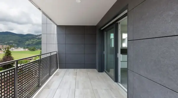 Nowoczesny balkon z jasnymi płytkami na podłodze i szarymi płytkami na ścianach. Widok na zielone wzgórza i budynki.