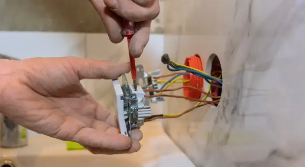 Ręce elektryka dokonują montażu gniazdka, przykręcając je do ściany. Widać kolorowe przewody wychodzące z puszki instalacyjnej.