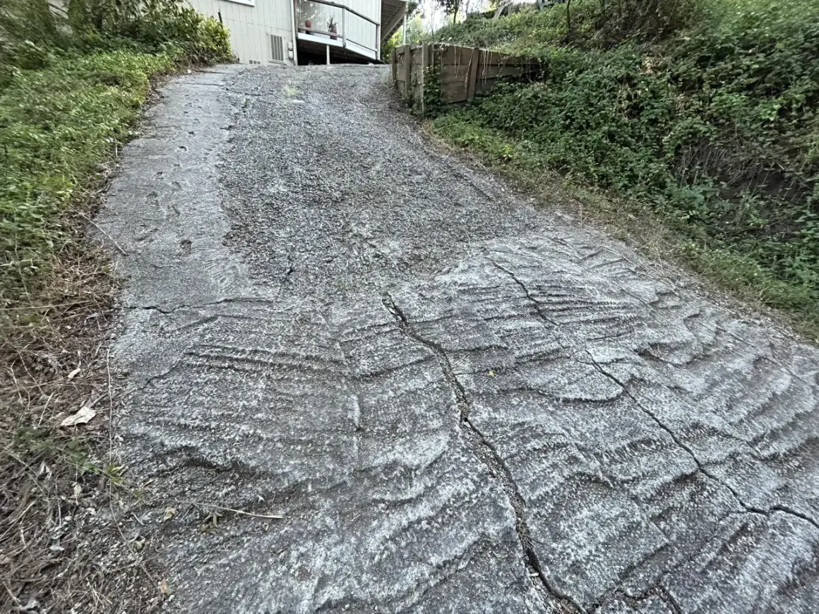 Wyboisty, wysoki podjazd do garażu, z widocznymi pęknięciami i nierównościami, otoczony zielenią.