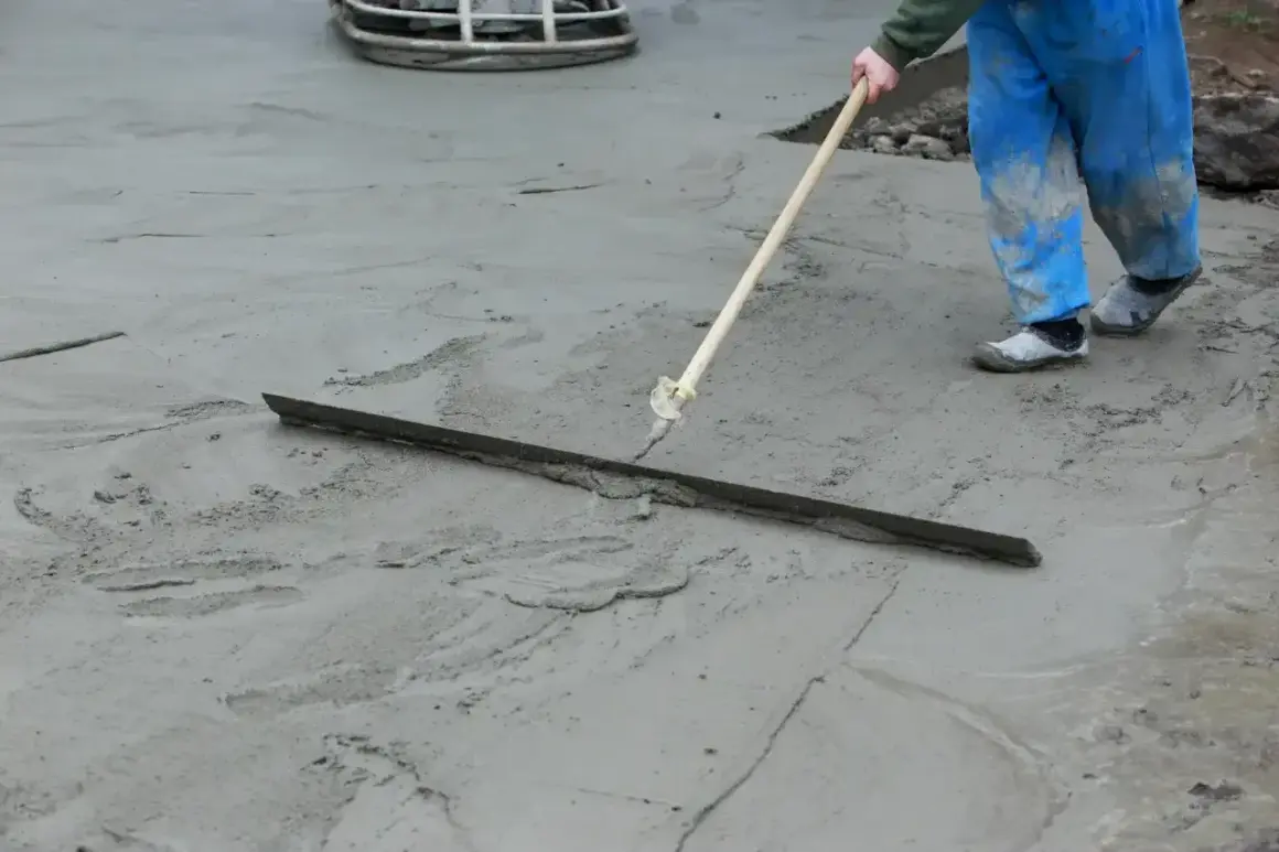 Pracownik w niebieskich ogrodniczkach wygładza świeży beton za pomocą łaty. Trwa wygrzewanie posadzki.