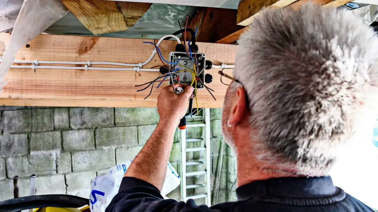Mężczyzna montuje instalację elektryczną w garażu. Widać skrzynkę z przewodami i narzędzia.