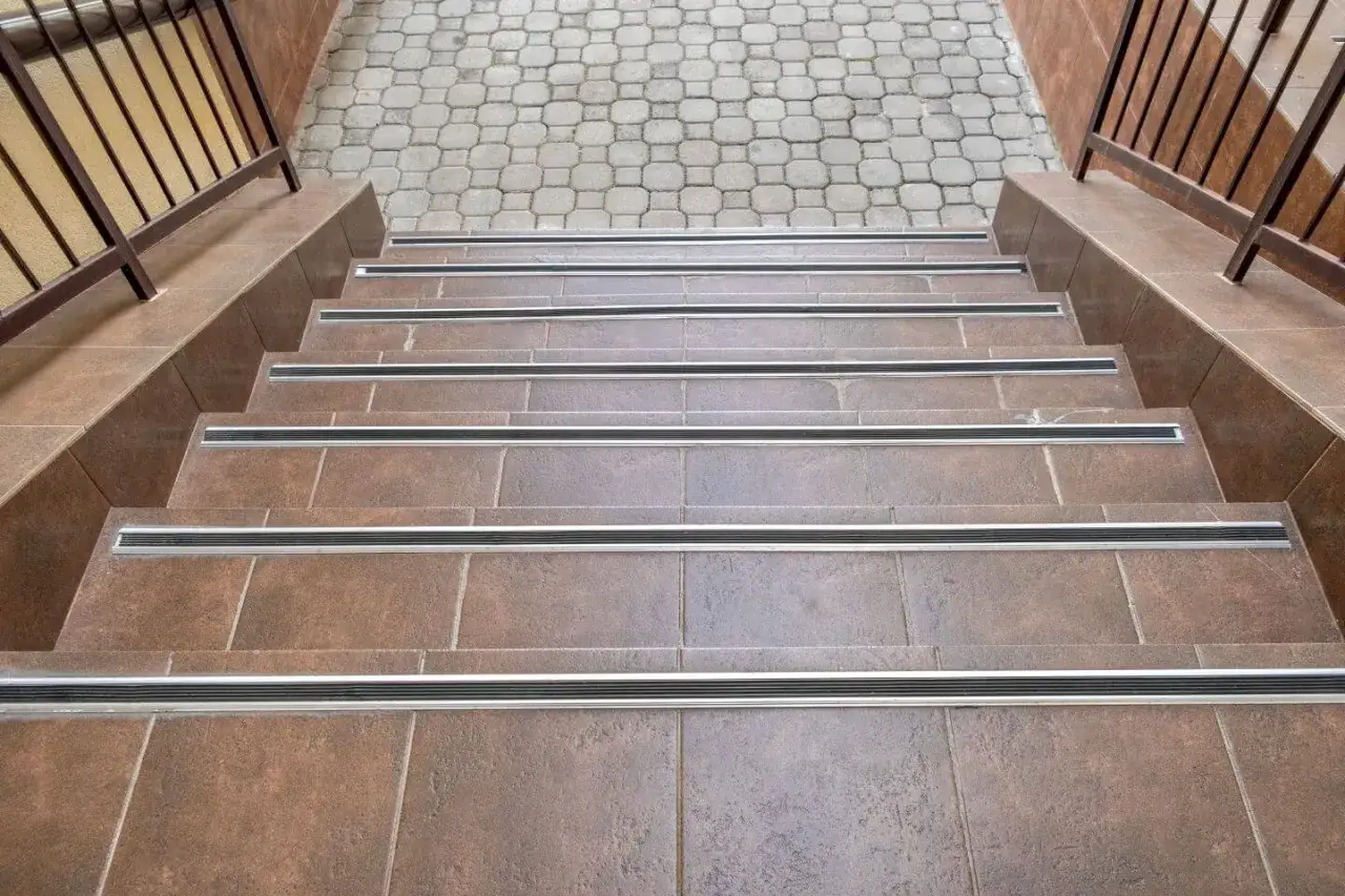 Schody z płytek ceramicznych, brązowe, z antypoślizgowymi listwami. U góry widać kostkę brukową i metalową poręcz.