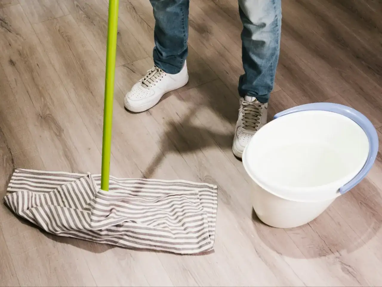 Osoba w jeansach i białych sneakersach przygotowuje się do mycia paneli. Obok stoi wiadro z wodą i mop.