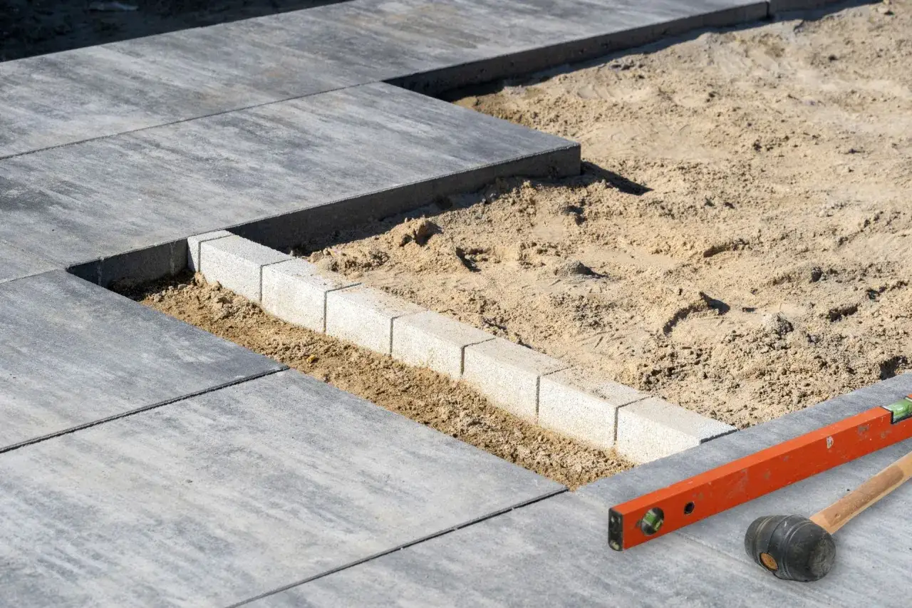 Budowa tarasu z betonu. Układanie płyt i obrzeży, w tle piasek i narzędzia: poziomica i gumowy młotek.