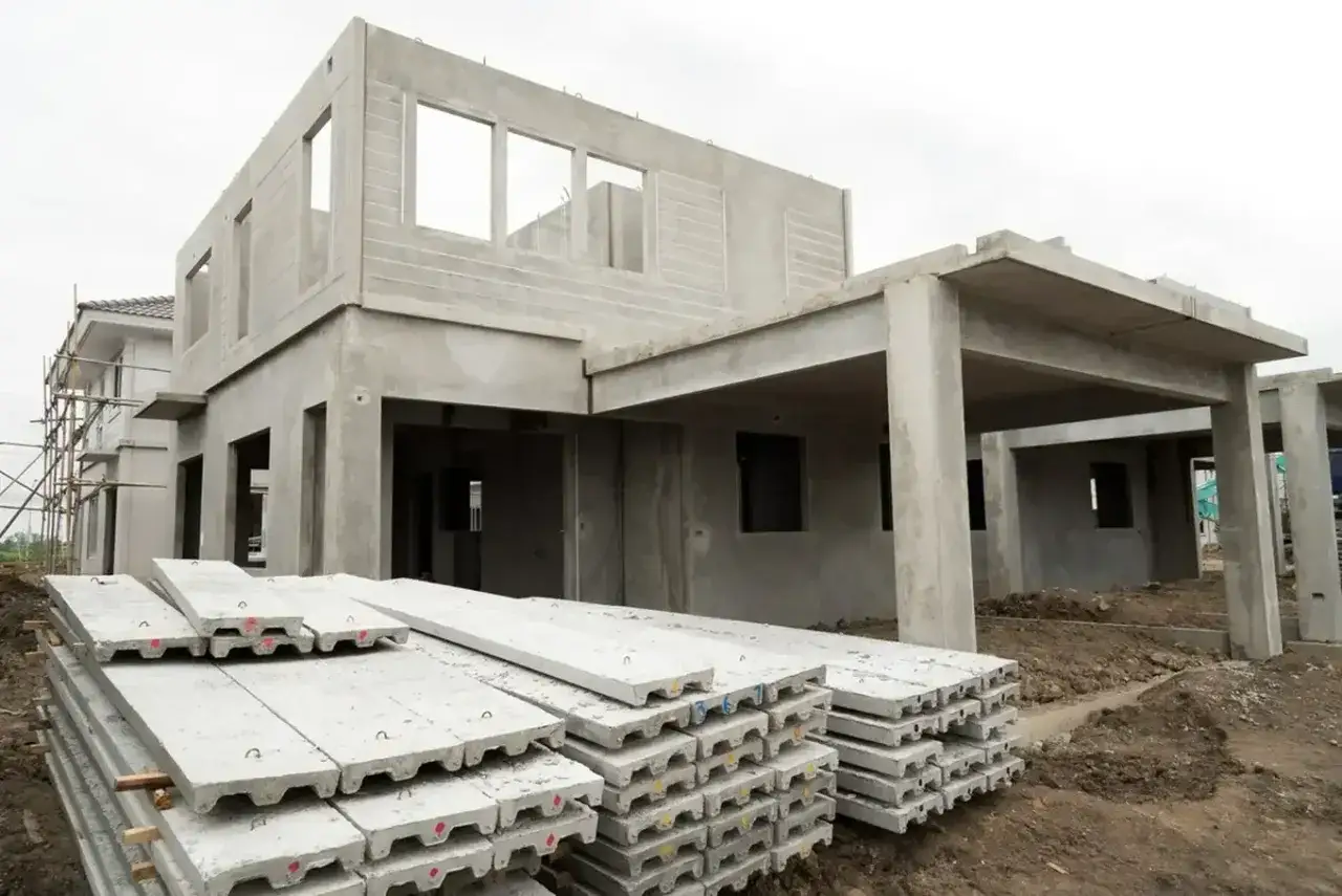 Budowa domu z prefabrykatów betonowych. Na pierwszym planie stosy płyt betonowych, w tle szary, niedokończony budynek z otworami okiennymi.