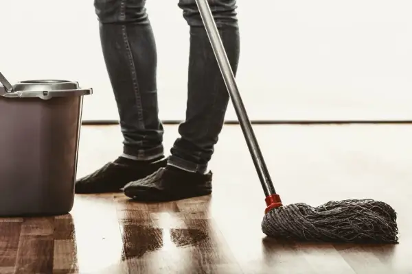 Osoba w jeansach i kapciach myje podłogę mopem, obok stoi wiadro. Naturalne czyszczenie octem.