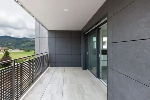Nowoczesny balkon z jasnymi płytkami na podłodze i szarymi płytkami na ścianach. Widok na zielone wzgórza i budynki.